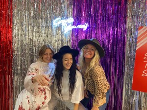 Three women posing in front of a neon sign that reads "Create Yourself"